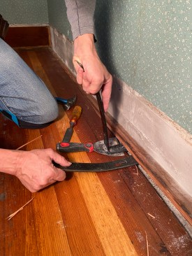 Once the tongue was split off, the last board could be pried free. Note that the author is using a wide trowel to spread the load of the flat bar in his left hand to prevent denting the floor surface. This is the bar he is using to exert the most force on to pry the board loose. The flat bar in his right hand is just holding the board up as he works along the length of the board, using the two flat bars in tandem.
