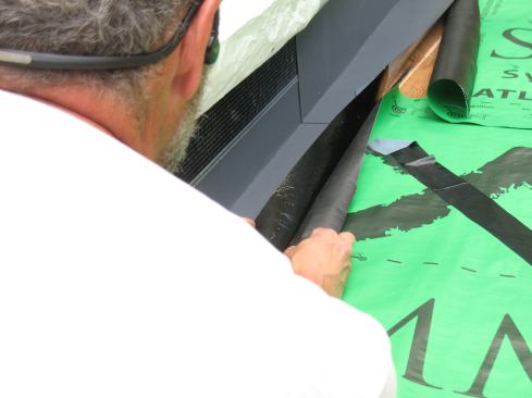 Because continuous flashing is less robust than step flashing, Mike backs it up with 12-inch flashing tape. He bonds 6 inches of the flashing tape to the wall and 4 inches onto the roof sheathing.