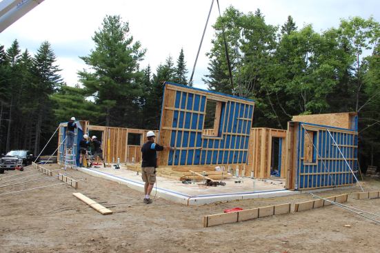 A crew member guides an end wall panel into place. At rear, the rest of the crew is plumbing and bracing the previously-placed panel.