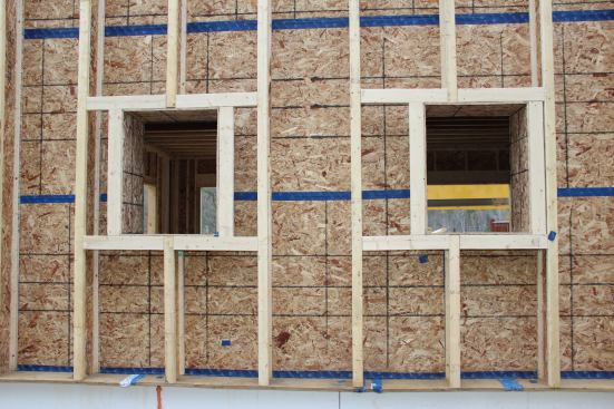 Completed window bucks, including blocking, for small windows on the building’s north wall. Tape over the OSB skin beneath the I-joists helps form the building’s air barrier. The corners of the window bucks are also taped, to integrate the window framing into the air barrier. An exterior drainage plane membrane, and strapping for the siding, will be applied next. Flanged windows will be fastened to the 2x4 blocking surrounding the OSB window bucks.