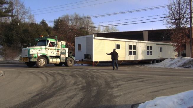 A tractor-trailer unit from Field and Sons (Grantham, New Hampshire) rolls out of the VerMod factory parking lot on a winter morning as Steve Davis stops traffic on Vermont Route 10 and serves as ground guide. The team has to wait until 8 AM, after school buses have finished their routes, to hit the highway with their police escort from the Windsor County Sheriff's Department. The VerMod units are considerably heavier than HUD-Code homes, and require a beefed-up trailer.