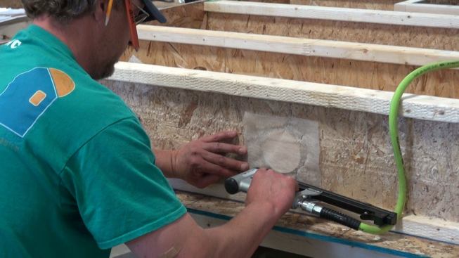 Stapling mesh screen over the pressure-relief hole in the wood I-joist at the edge of the wall panel. This opening turned out to be necessary because the I-joist cavities, once covered with taped fabric, are air-tight enough to create a back-pressure the can impede the operation of the insulation blower unless relief is provided.