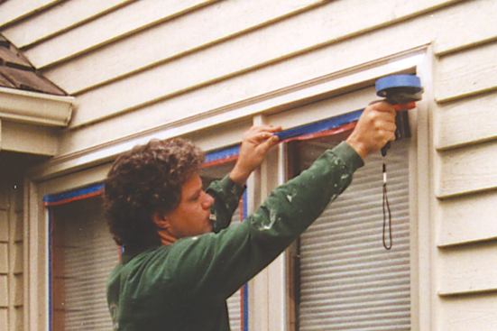 Spraying is considerably faster than brushing when windows, siding, and trim are being painted at the same time. The author begins by using a dual-tack tape dispenser to mask the window glass.