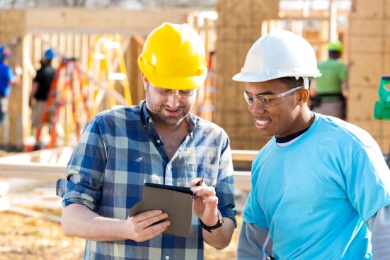Construction foreman discusses building plans with construction worker. The foreman is using a digital tablet. Construction woerkers are building new home in the background.