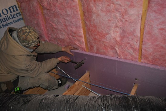 Carpenters notch and fit 2-inch XPS insulation board over the wall studs, fastening the board with nails and washers as they compress the R-19 batts into the wall framing cavities.