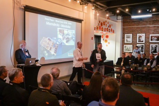 NADRA president Matt Breyer congratulates Joel Maendel of Maendecks, winner of the third-place honor in the $25,000 to $50,000 wood deck category in NADRA's 2019 national deck competition. Master of ceremonies Bobby Parks (at the podium) looks on.