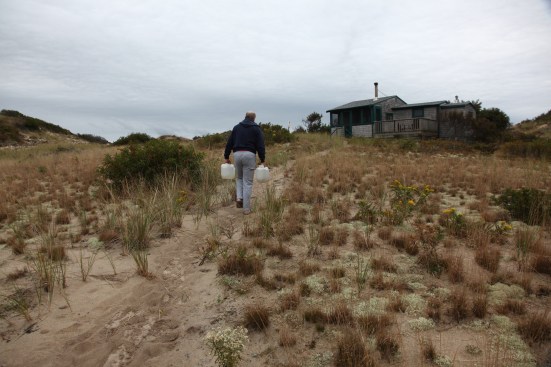 After pumping and filling the jugs, the author carries the water uphill back to the shack.