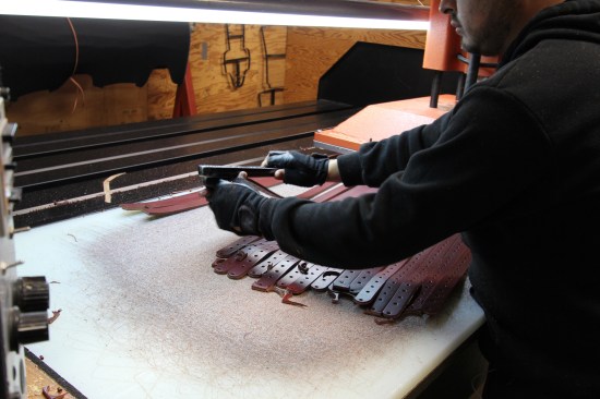 The operator peels the finished piece out of the die after the press punched it through. These suspender straps will pass through buckles—hence the many holes for adjusting their length.