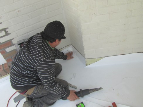 A worker presses a pre-shaped corner piece into place at an inside corner joint in a TPO roof. In his right hand is the heat-welding gun, used to melt both pieces in the joint before pressing them together to form a durable seal.