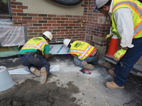 Flashing membrane is applied to the wall as a backing for the new stonework, then a base course of stone is set against the flashed wall. Another through-wall flashing is installed before upper courses of stone are laid (leaving weeps that allow the wall to drain).