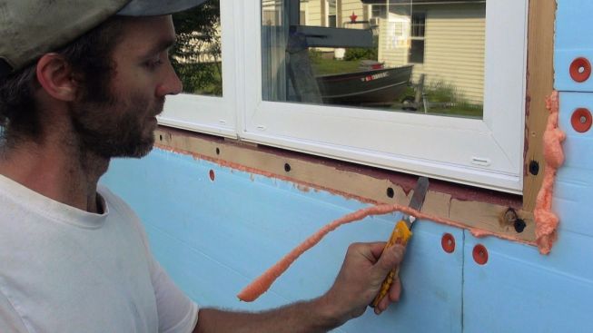 Because the existing walls were out of plumb, windows were installed in the center of the window openings to enable crews to split the difference while plumbing the window units. Then, Dow Styrofoam insulation was applied to the wall exteriors. Here, David Kelly trims off excess injected foam used to fill and seal the gap between the XPS foam and the furred window opening.