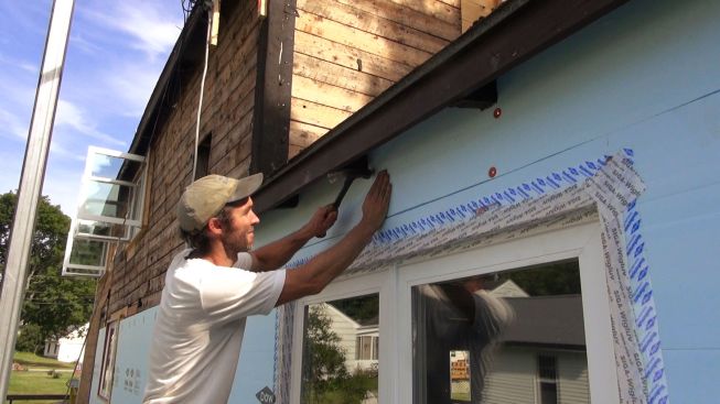 Kelly fastens the piece of foam in place with a few cap nails. The cap nails serve to hold the piece until strapping is applied over the foam and screwed to the house sheathing for a secure attachment.