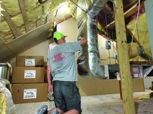 An installer for Air Rite By Design tapes insulation on a duct. Even though the ducts are within the conditioned envelope, they still must be insulated. At right is a multi-position air handler for a variable refrigerant flow (VRF) system. The air handler is suspended from the roof framing with threaded rod and vibration-damping couplings.