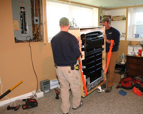 Electricians Scott Millette and Brian Ritz of Peck Electric (South Burlington, Vermont) carry a Tesla Powerwall, still in its crate. The unit weighs 214 lbs.