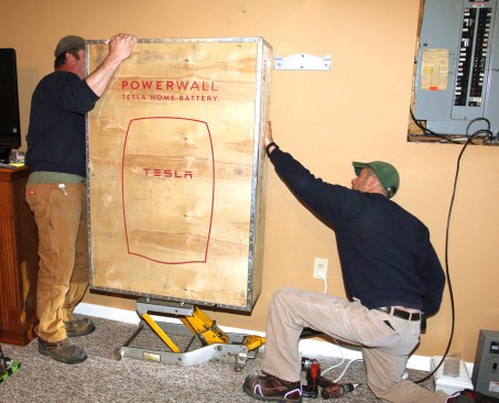 Using a motorcycle jack, electricians Ritz and Millette position the Powerwall battery into place. The battery is hung from clips on the mounting bracket.