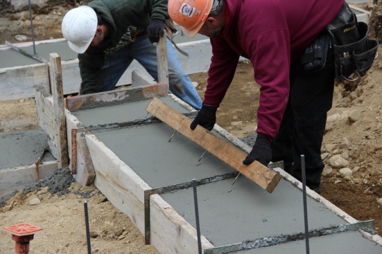 A concrete mason marks rebar locations in the freshly placed concrete footing using a jig made by inserting screws into a 2x4.