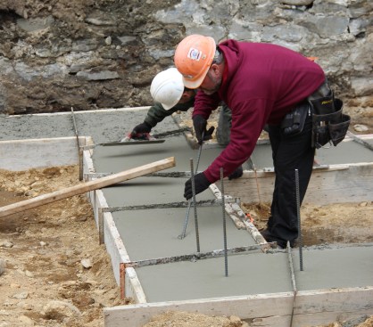 A concrete mason places half-inch rebar bent at a 90-deree angle into the freshly placed concrete at the pre-designated spacing. He hooks the rebar underneath the horizontal rebar that was placed 