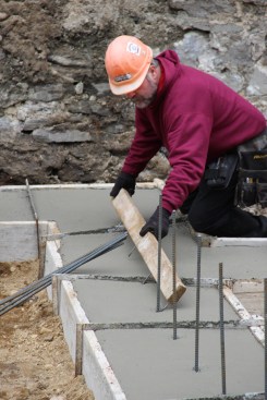 A concrete mason uses a jig made of screws set into a 2x4 to mark the freshly poured footing at two-foot on-center spacing for vertical steel rebar, which will tie the footing to the foundation wall.