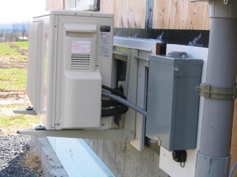 The outside compressor is mounted on a wall bracket, with the sub-panel on the right. The line set and 4-conductor wire pass through a sleeve to the interior. Before bracket installation, the author applied bituminous membrane to the sheathing and exterior trim to accommodate the finish siding.