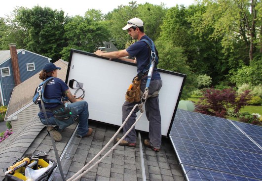 Technicians for Revision Energy install PV panels for a grid-tied system on a Portland, Maine, roof in 2014. A panel array this size might supply all the power a house needs on a net annual basis, but still requires a grid connection to balance surplus summer production against nighttime and winter power needs.