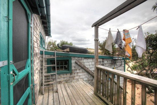 Steps lead to a small deck for the back entry into the dune shack. Clothes drying on the line indicate that the shack is occupied. The short ladder is for filling the black plastic container that acts as a solar water heater.
