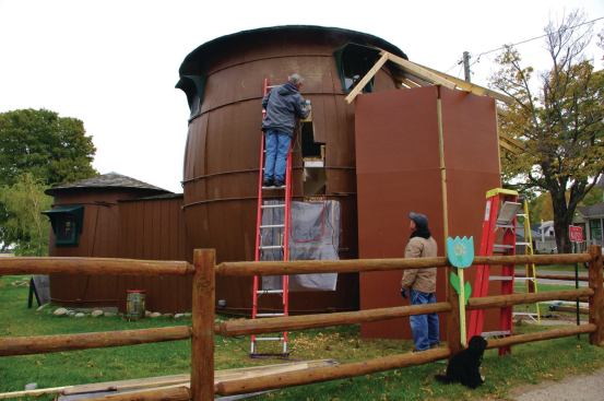 Boatbuilder George Powell cuts a scarf joint in an existing stave to splice in a steam-bent replacement. The smaller “kitchen barrel” at left was in better shape, thanks to a roof overhang large enough to direct water from the eaves beyond the outward-bulging walls. The temporary structure at right protects the open doorway.