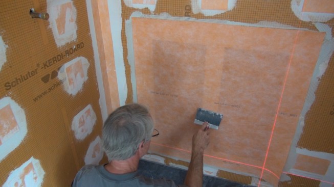 Boucher uses a six-inch drywall knife with the corners ground down to press the membrane into the mortar. He can see well enough through the membrane to visually check that the quarter-inch grooves in the mortar have been fully smoothed down, indicating a full mortar bond with the membrane.