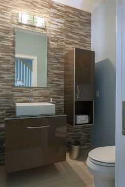The new bathroom features a wall-mounted vanity with highly polished laminate bronze finish, a glass counter top, a motion activated wall-mounted sink, and a primary wall covered with mixed linear glass mosaic tile.