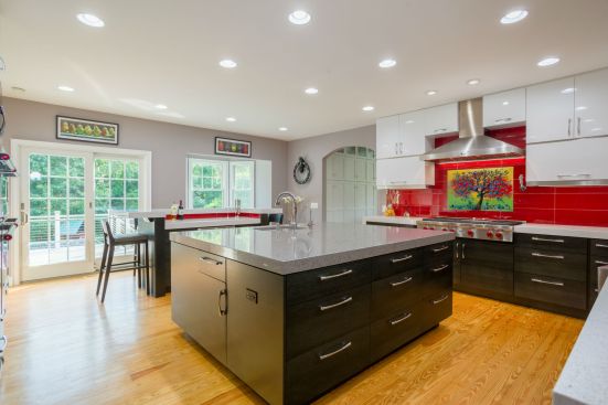 Clean white cabinets above complement the stained black, quarter-sawn horizontal grain oak that lines the kitchen at its base, providing the Tuxedo Kitchen theme. In place of the original bay window, sliding patio doors were used to allow more room for a secondary, double-tier seating/work island topped with 3-inch quartz. The doors provide an added touch of natural light to a room the clients requested to include real outdoor lights controlled by dimmers.