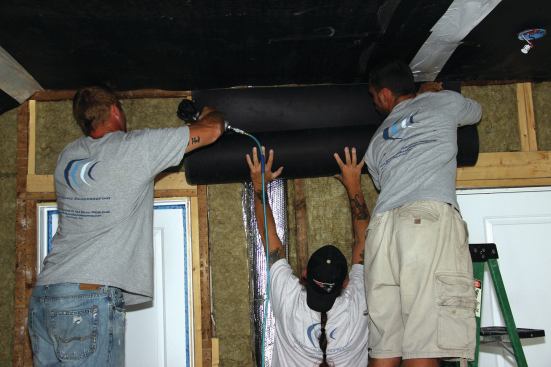 The crew installs a "mass-loaded vinyl" (MLV) membrane over stud cavities filled with sound-blocking insulation such as Roxul rock wool or QuietInsul. 