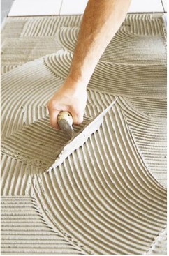 After applying the setting adhesive with the straight edge and bottom of the trowel, the tilesetter combs the setting bed with the trowel held at a 45-degree angle.