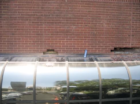 The offices under this curved glass roof had been getting wet for years. The drops of water usually came down days after the rain — not surprising, given the height of the brick wall above, a likely water reservoir. I started with some controlled testing and discovered that water was showing up inside about 10 feet away from an area where I could see a lap in the metal through-wall flashing outside.