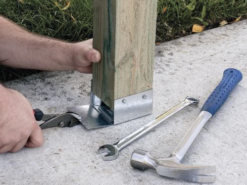 Most post bases fasten to a bolt that’s cast or epoxied into the concrete footing. Once the post is located and the nut tightened, the base is nailed to the post.