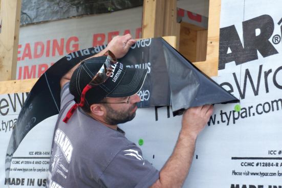 Start at the bottom of the window opening. Adhere a flashing strip to the sheathing and housewrap as shown.