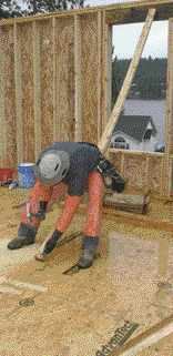 A carpenter nails the brace to the wall, then springs it down by sitting on it before nailing it to the deck.